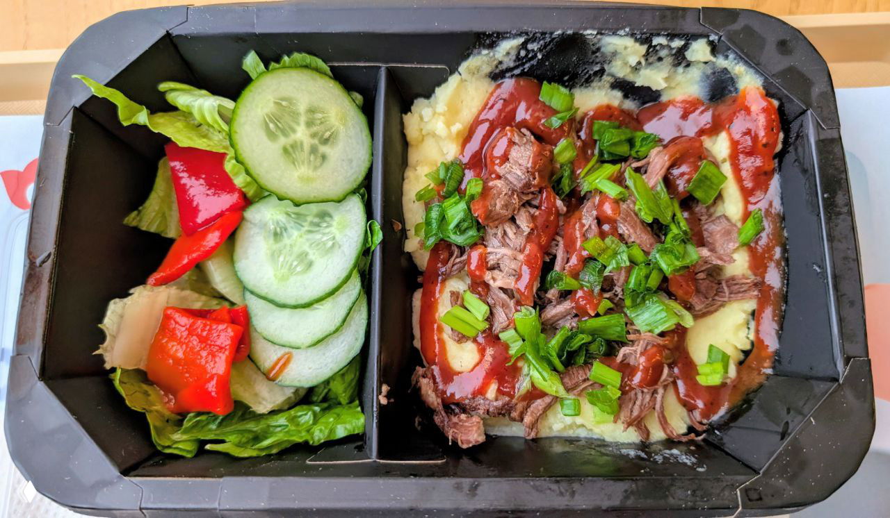 Контейнер с блюдом: мясо под соусом и гарнир, рядом свежий салат — пример обеда в Казани.