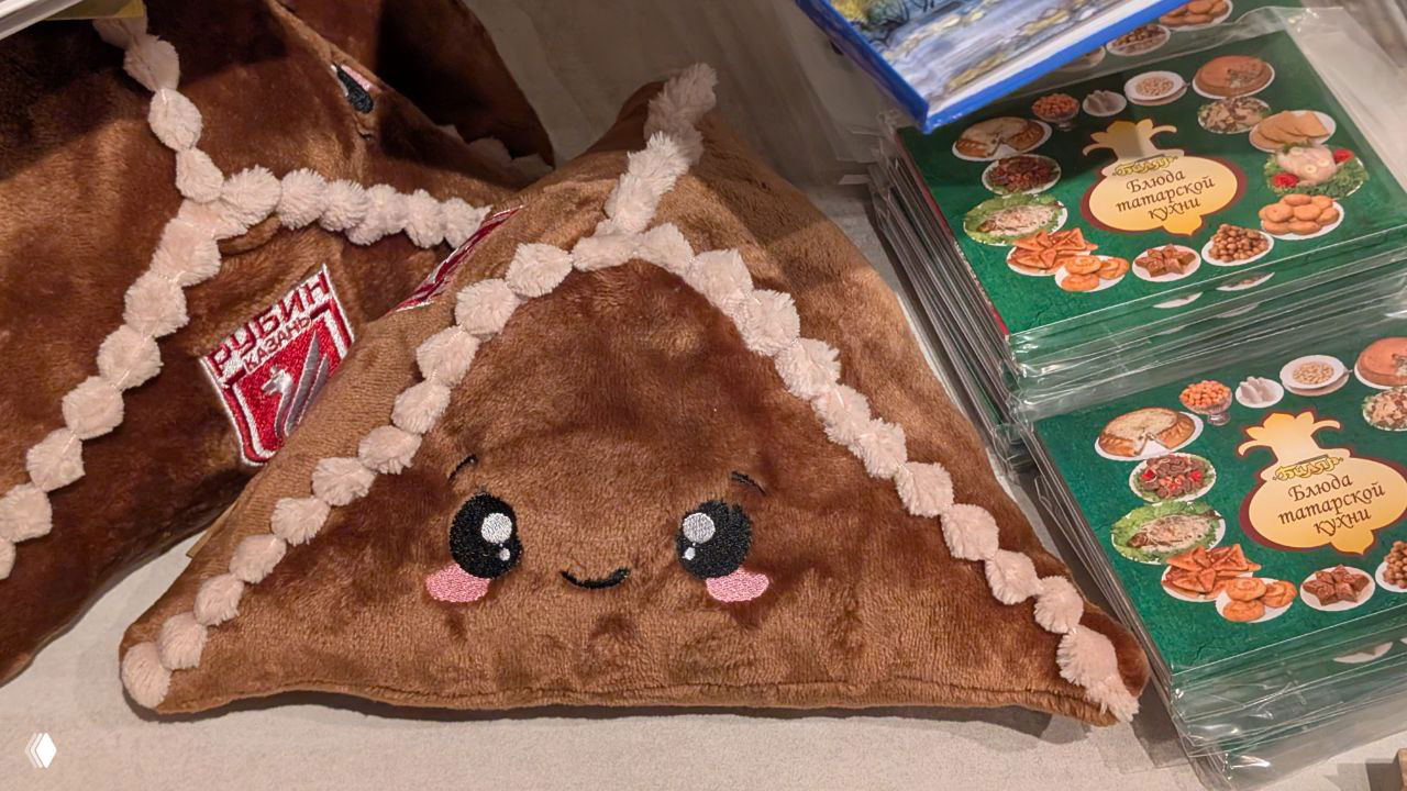 Плюшевая подушка в форме эчпочмака на сувенирном столе рядом с книгами о татарской кухне — сувениры из Казани.
