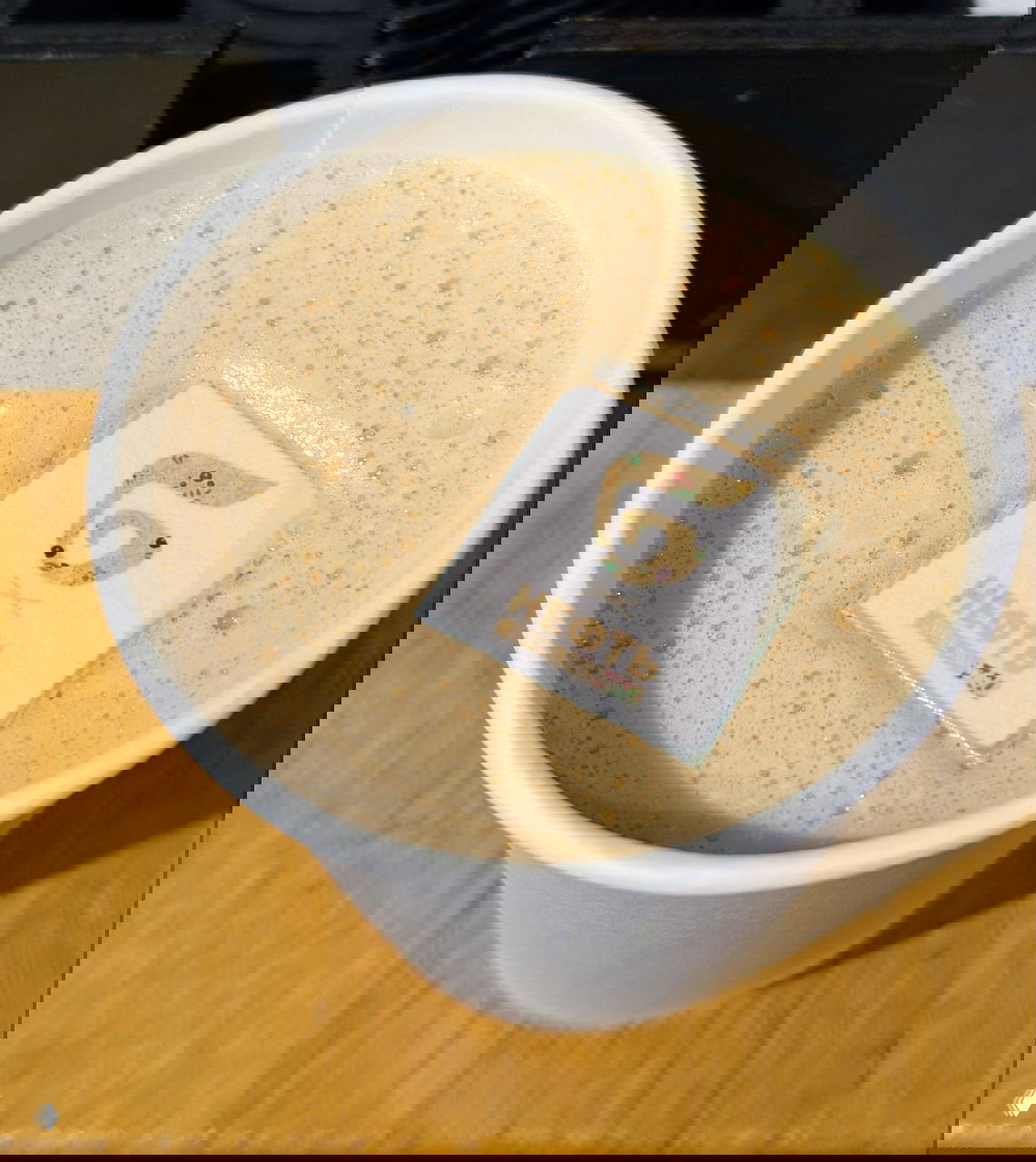 Бумажный стакан с пенистым авторским напитком кофейни «Нефть», сверху декоративный сахарный квадратик с логотипом заведения.