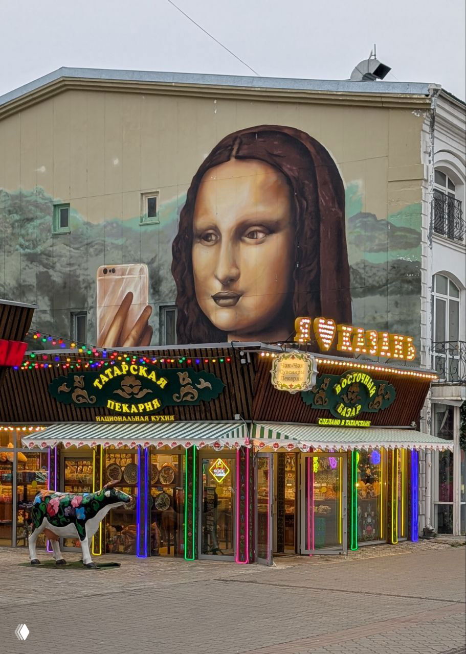 Яркий мурал с изображением Моны Лизы, держащей смартфон, над ярко освещённой витриной татарской пекарни на пешеходной улице Баумана.