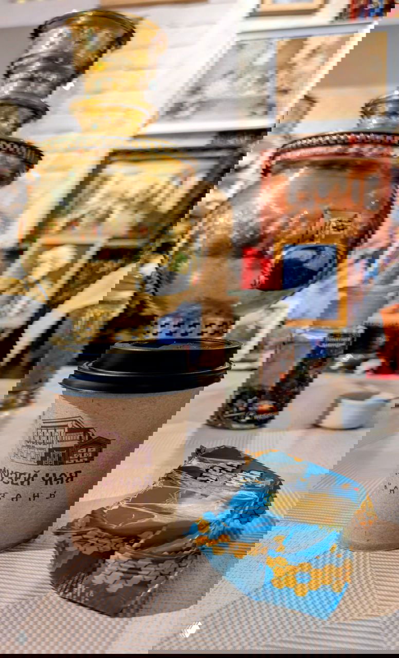 Чашка на столе и бумажный стакан с логотипом Музея чая Казань на фоне большого самовара и интерьерных деталей музея