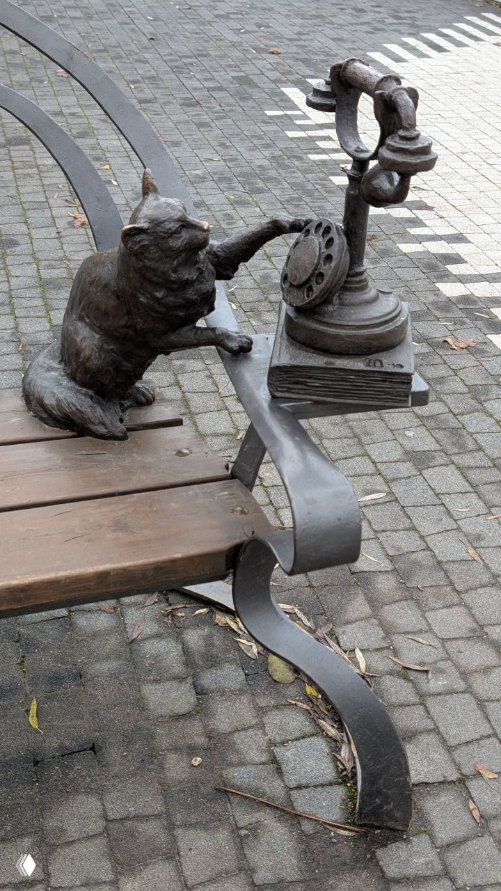 Бронзовая кошка Ю-ю на скамье тянется к старинному дисковому телефону — часть скульптурной группы, связывающей литературу и бытовую деталь.