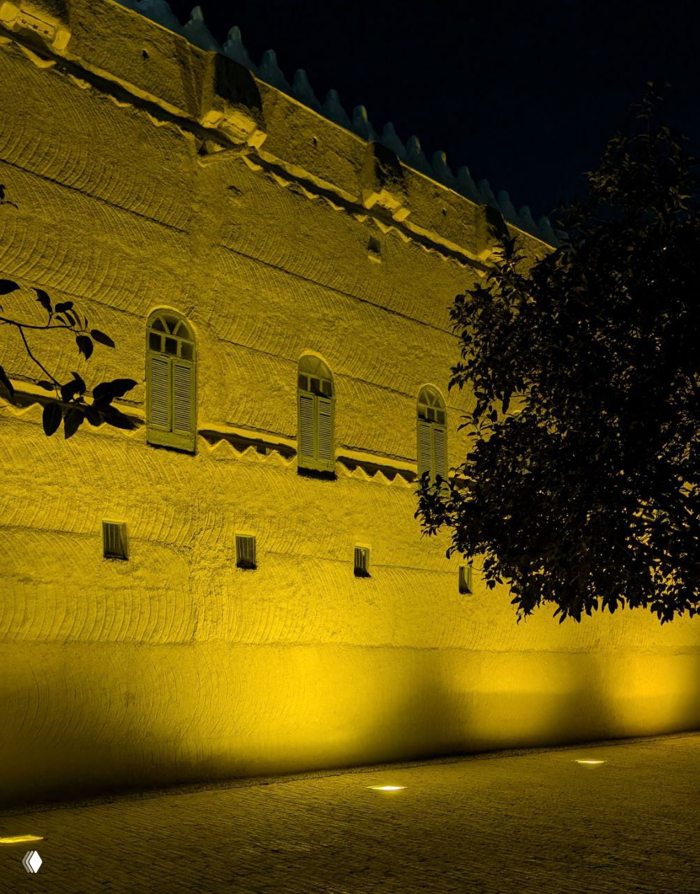 Подсвеченный фасад традиционного арабского здания ночью: узкие окна, рельефная текстура стены и силуэт дерева на переднем плане.