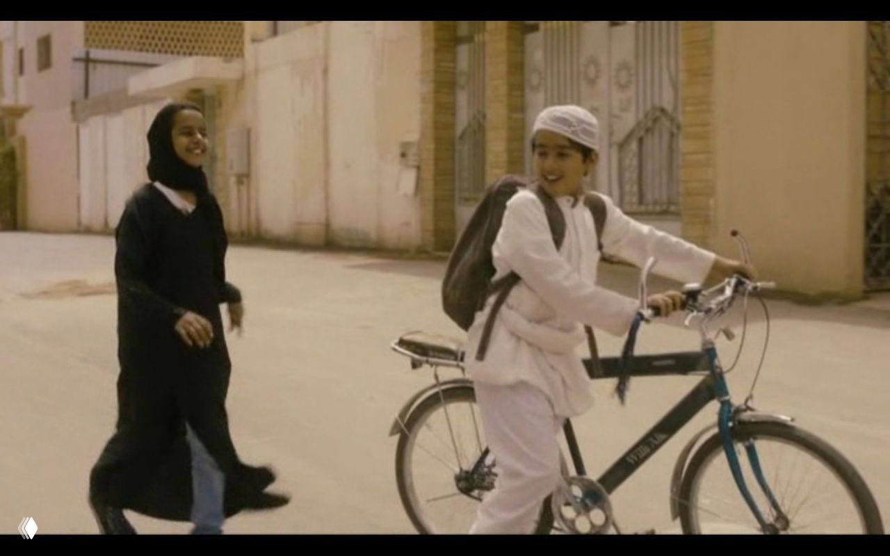 Уличный кадр из «Ваджда»: девочка радостно едет на велосипеде рядом с подругой в абайе — образ желания свободы на фоне традиций.