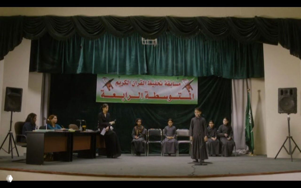Сцена школьного конкурса из «Ваджда»: сцена, участники и баннер на арабском — сюжетный эпизод с конкурсом по чтению Корана и призом в 1000 риялов.