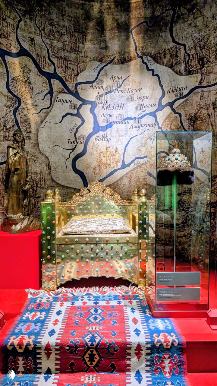 Трон, ковёр и корона в музейной витрине, оформленные как часть экспозиции Казанского ханства с картой на заднем плане и витринной подсветкой.