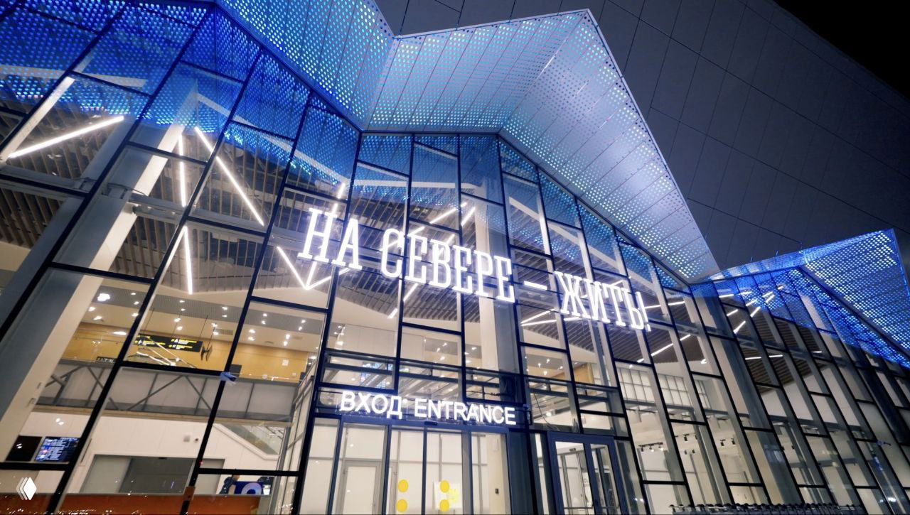 Фасад нового терминала аэропорта ночью с подсветкой и надписью «На Севере — жить», стеклянный вход и яркие LED-панели.