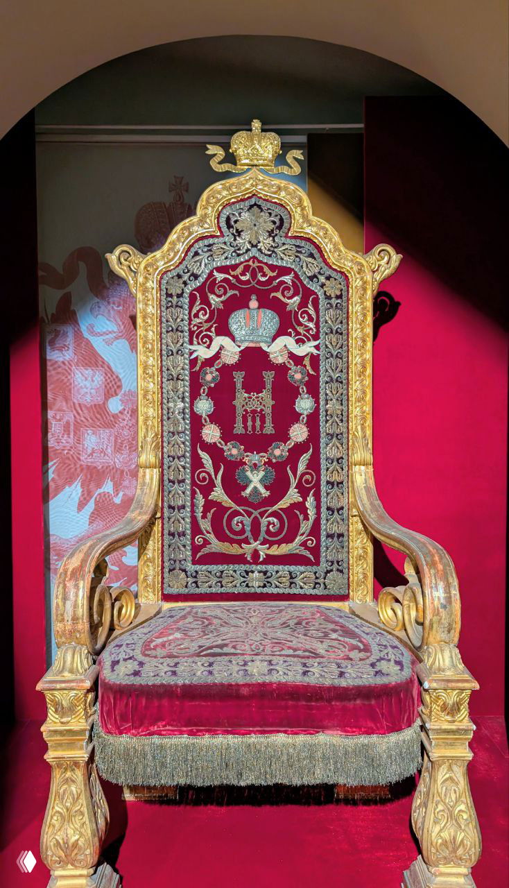 Бархатное тронное кресло с вышитой монограммой и короной, резная позолоченная спинка и подлокотники, выставлено на красном фоне в Троицкой церкви Гатчины