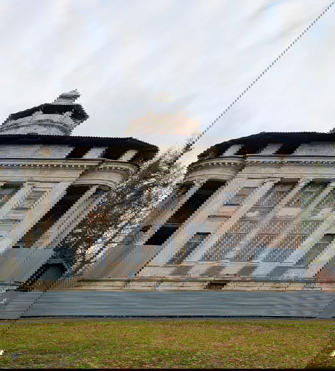 Фасад главного дома усадьбы Тайцы с бельведером и колоннадами за металлическим забором, вид с передней стороны в пасмурный день.