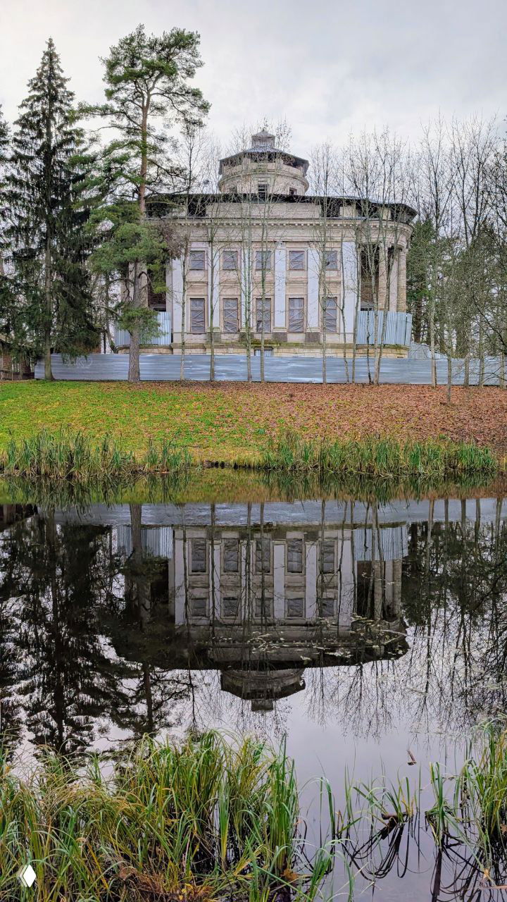 Отражение здания усадьбы Тайцы в тихой заводи паркового пруда; по берегу осенние травы и голые деревья, пасмурная погода.