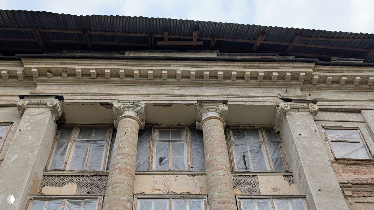 Крупный план фасадных колонн с коринфскими капителями и частично заклеенными окнами — следы разрушения и временной защиты.
