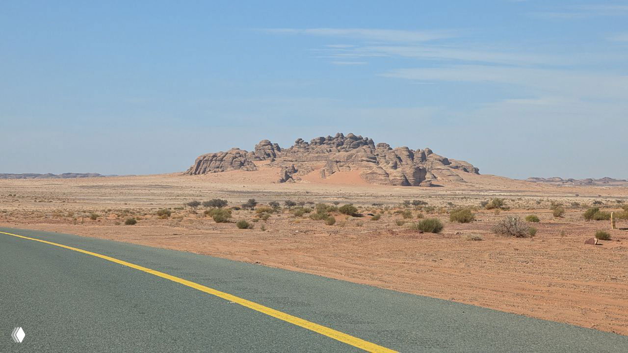 Пустынная шоссейная дорога в Саудовской Аравии: асфальтированная трасса вдоль ровного пустынного ландшафта и одинокая каменная скала на горизонте.