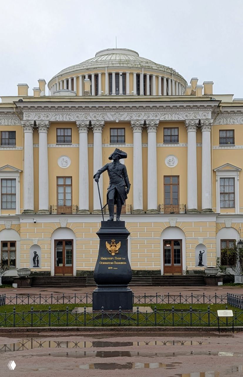 Фасад Павловского дворца с памятником Павлу I на переднем плане: классический фасад, колоннада и постамент со статуей перед входом.