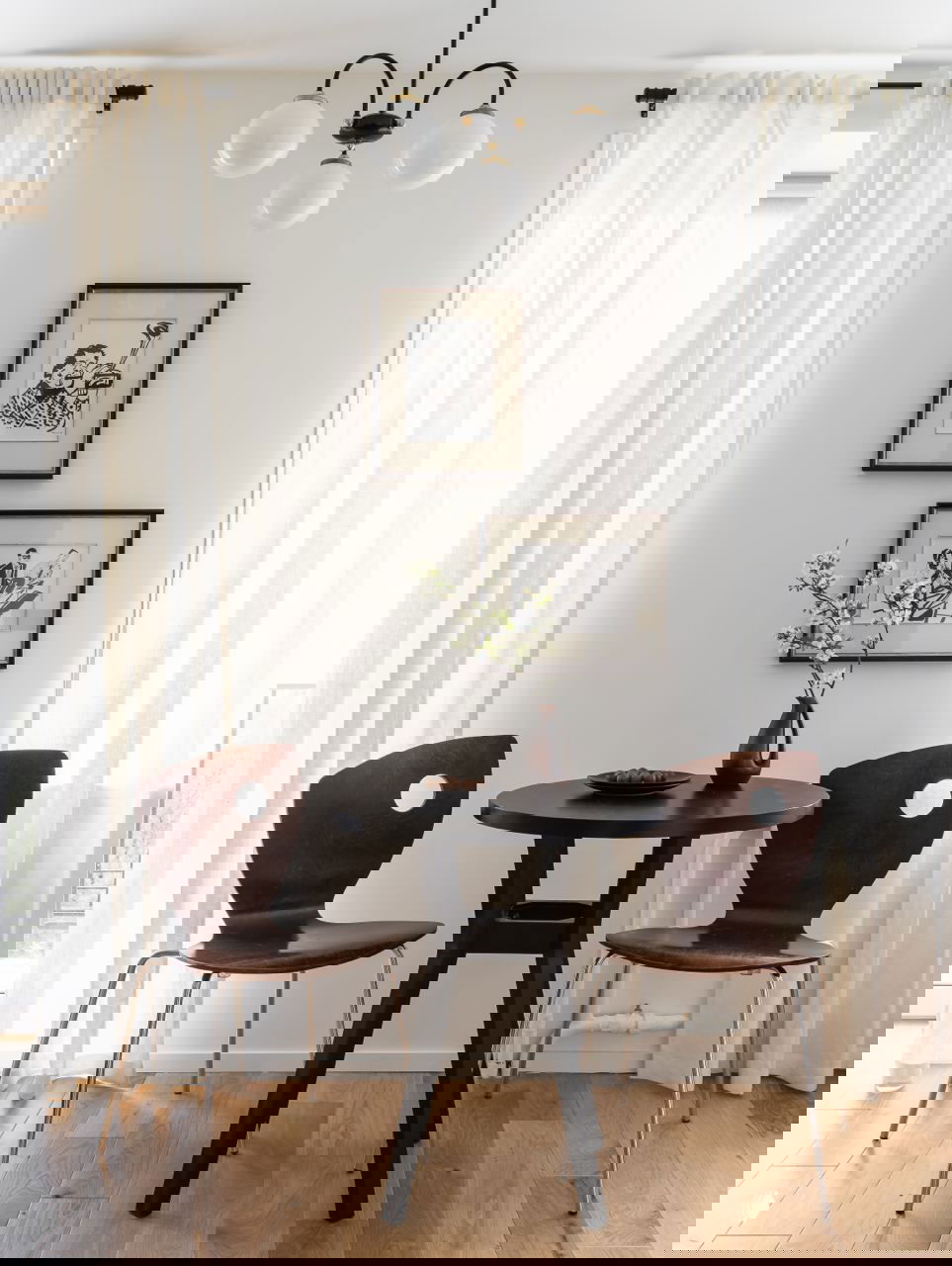 Небольшая обеденная зона в хрущевке: круглый тёмный стол, две ретро‑стула с вырезом, светлая штора и подвесной шарообразный светильник над столом.