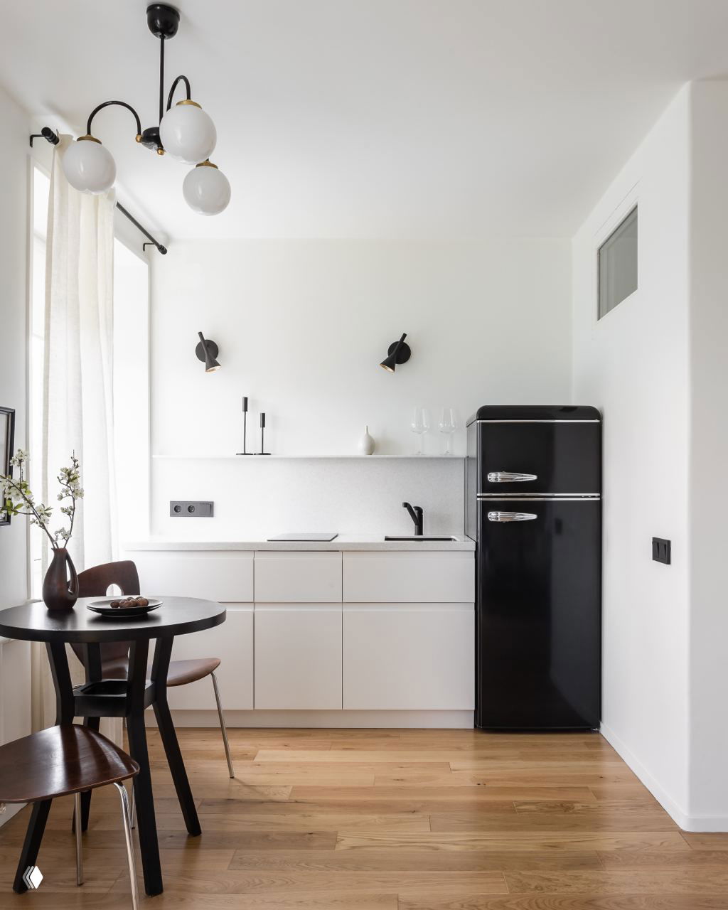 Кухонная ниша в маленькой квартире: белые лаконичные шкафы, тёмный кран и ретро‑чёрный холодильник на фоне светлых стен и деревянного пола.