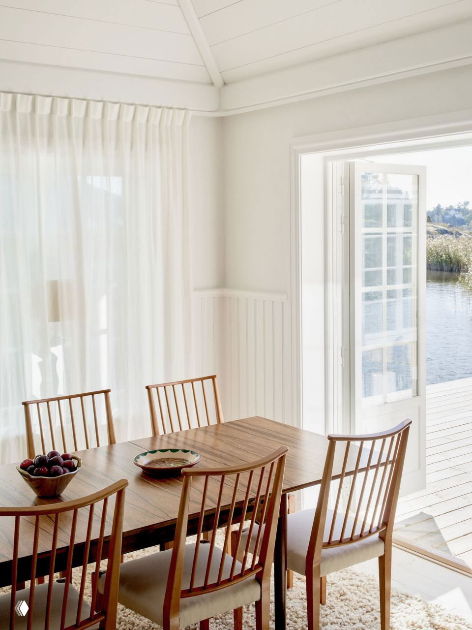 Светлая столовая с деревянным обеденным столом и стульями у распахнутой двери: вид на берег моря, мягкий текстиль и солнечный полутон в интерьере дома-эллинга.