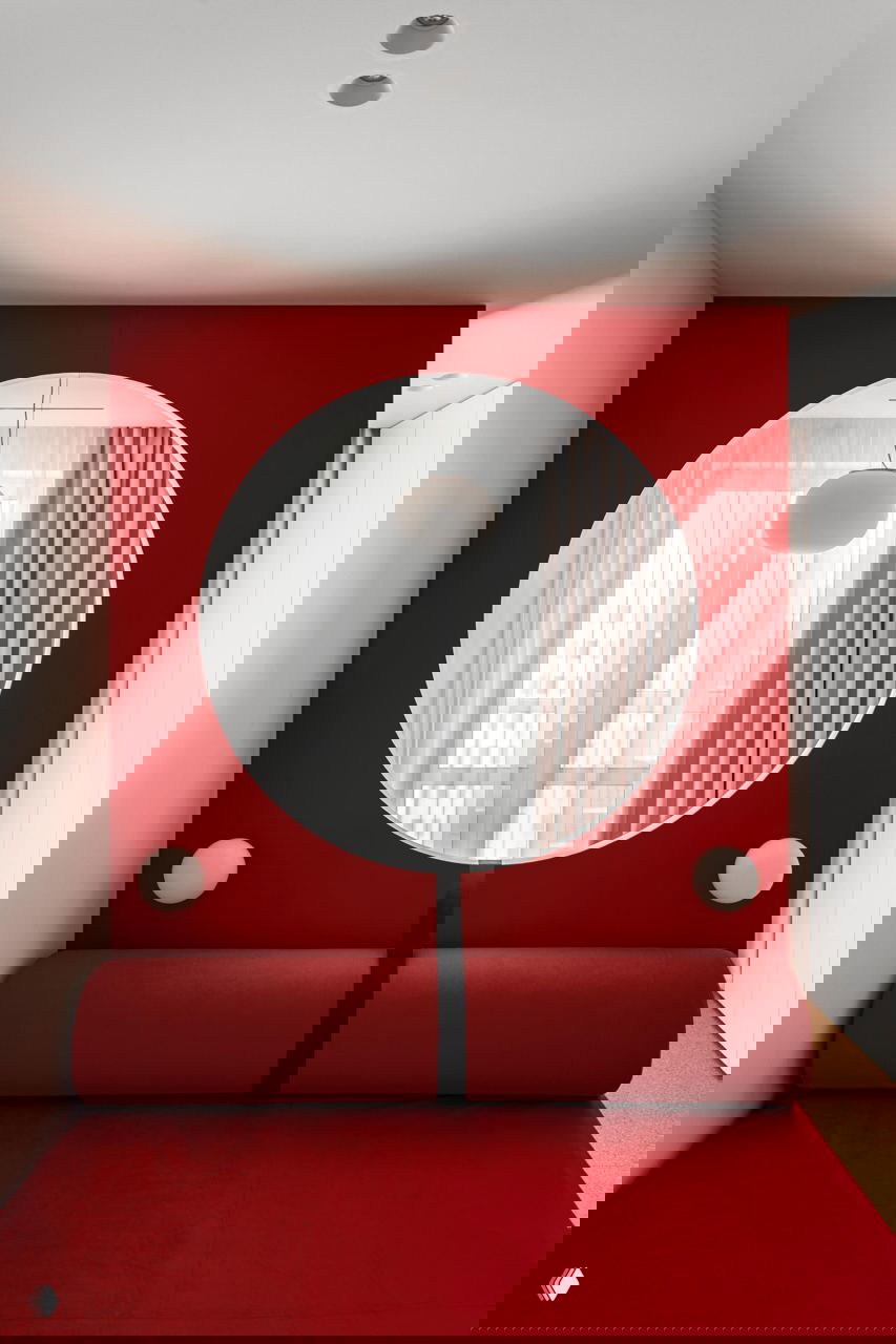 Ниша с большим круглым отверстием в красной стене, через которую видно окно с подвесным светильником; внизу роликовый диван и яркие текстуры.