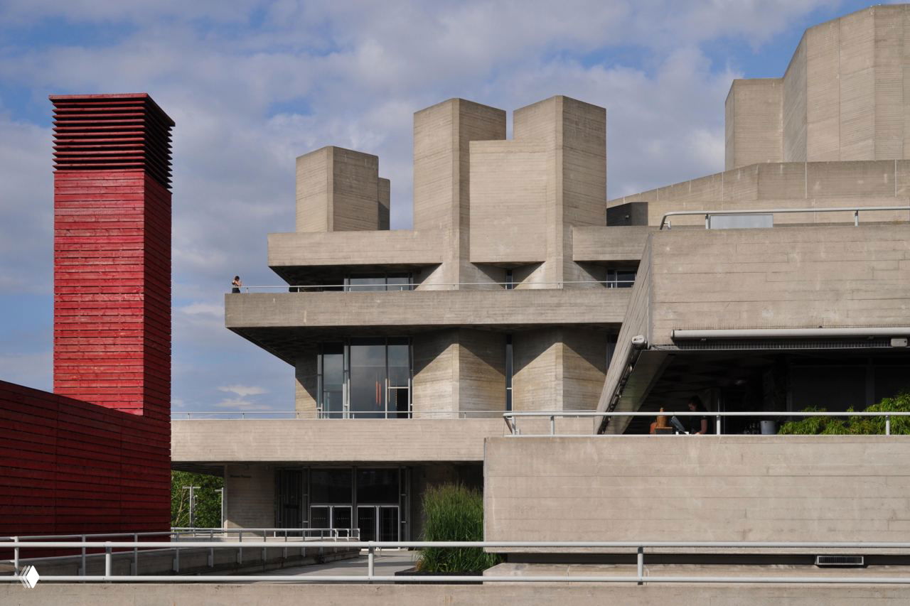 Фрагмент бруталистского бетонного комплекса с массивными кубическими объёмами, террасами и вертикальной красной шахтой — акцент на монументальности и фактуре бетона.