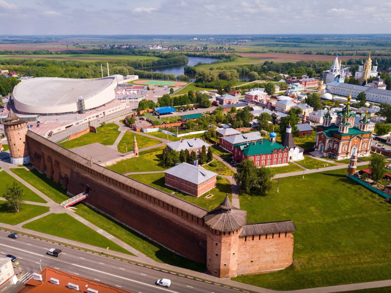 Панорамное аэровидение Коломны с крупной кирпичной стеной Коломенского кремля, башнями, зелёными полями и рекой на дальнем плане.