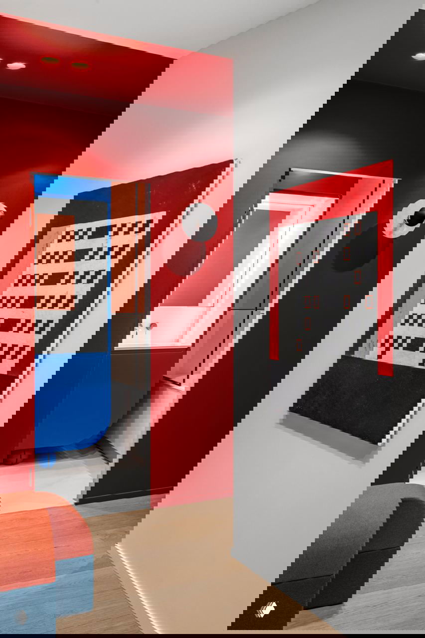 Красный коридор с проёмом в ванную, на белой стене висит графическое полотно; на полу — деревянный паркет, видна синяя сантехника за дверью.