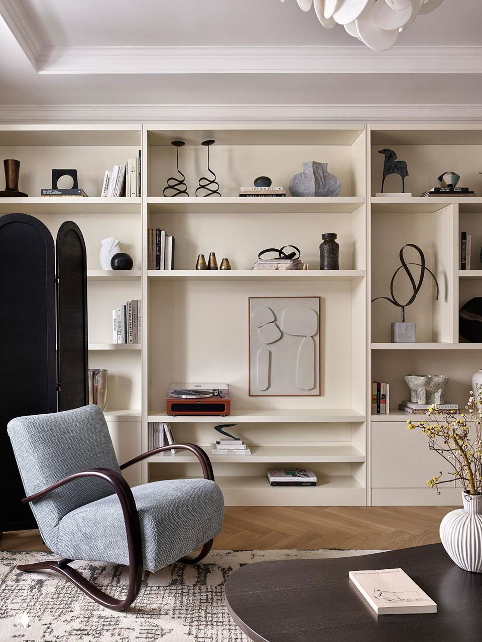 Гостиная с мягким креслом и открытыми бежевыми полками, в интерьере заметны чёрные декоративные акценты, виниловый проигрыватель и деревянный паркет.
