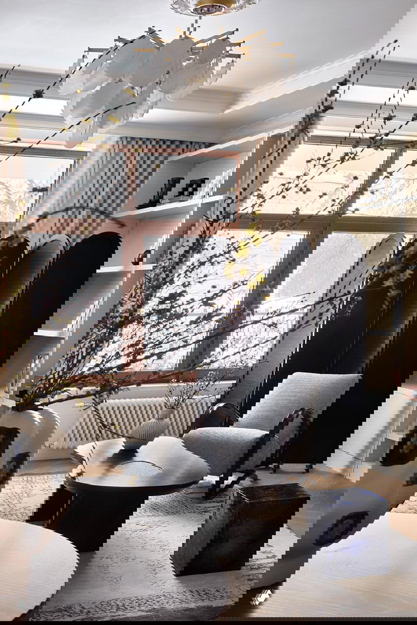 Уютная зона у окна: кресло, кофейный столик, декоративная ширма и карнизное окно с видом на город, в кадре видно весенние ветви в вазе на переднем плане.