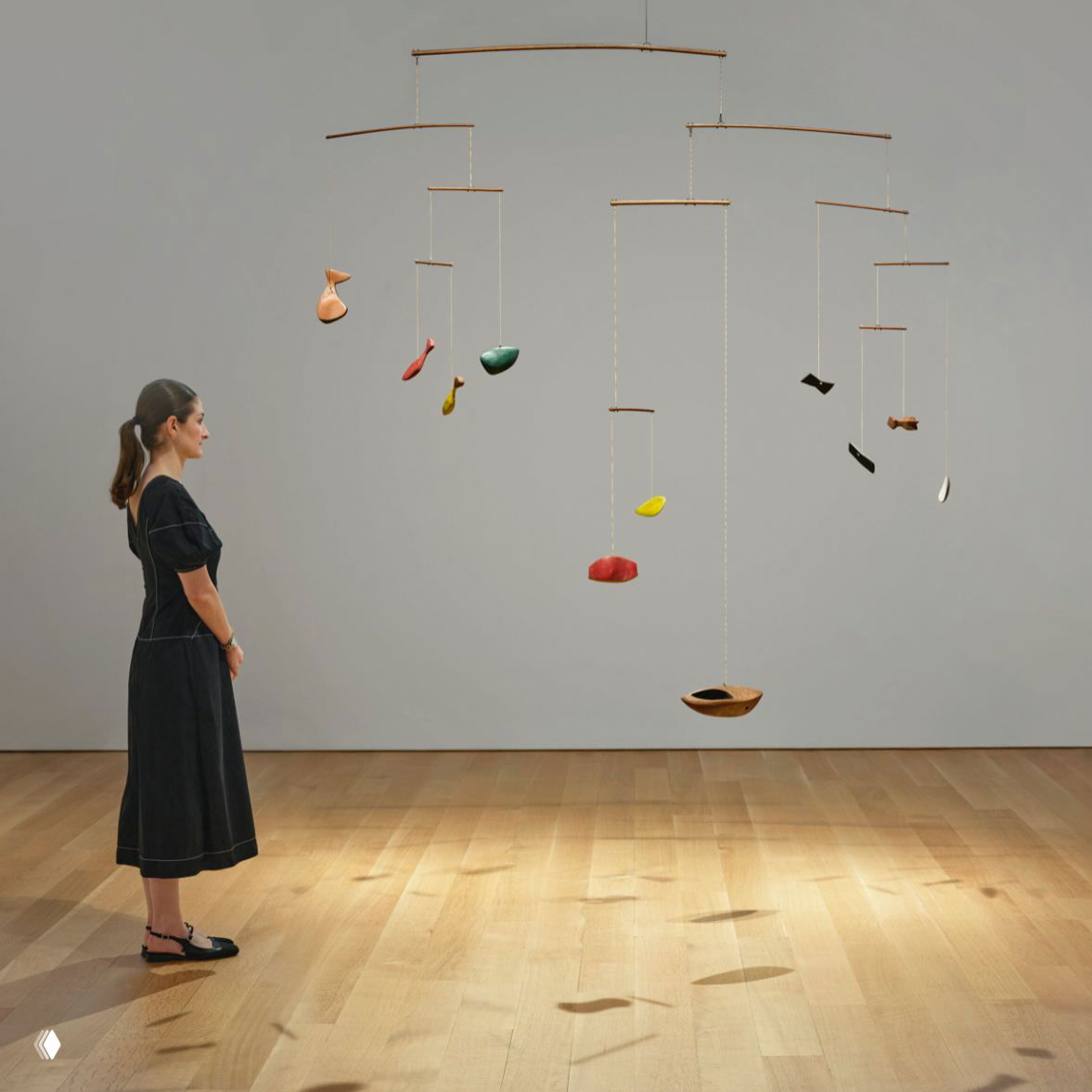 Большой кинетический мобиль Александра Колдера «Раскрашенное дерево» на нейтральном фоне в зале галереи; рядом стоит женщина для визуального масштаба экспоната.