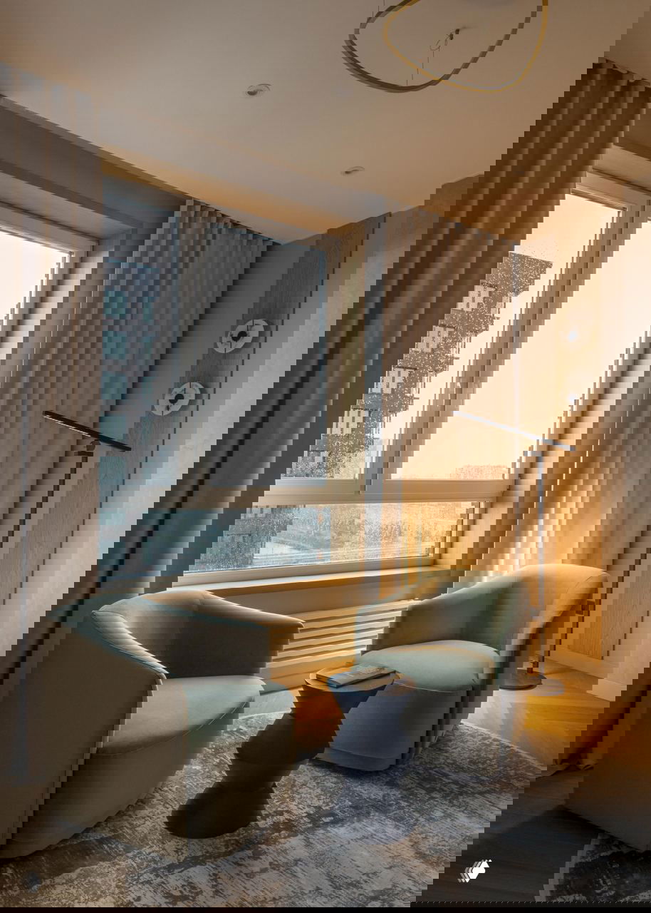 Уютная читающая зона у окна: пара оливковых кресел, торшер и низкий столик на фоне крупного окна с видом на город и тёплым освещением.