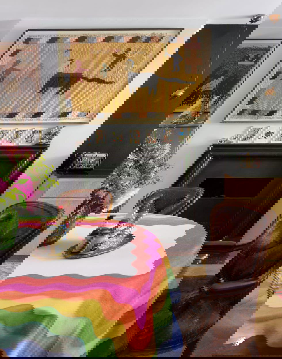 Столовая зона с круглым столом в радужной скатерти, пара кожаных кресел и крупное декоративное полотно над темным камином — насыщенные цвета и винтажные детали.