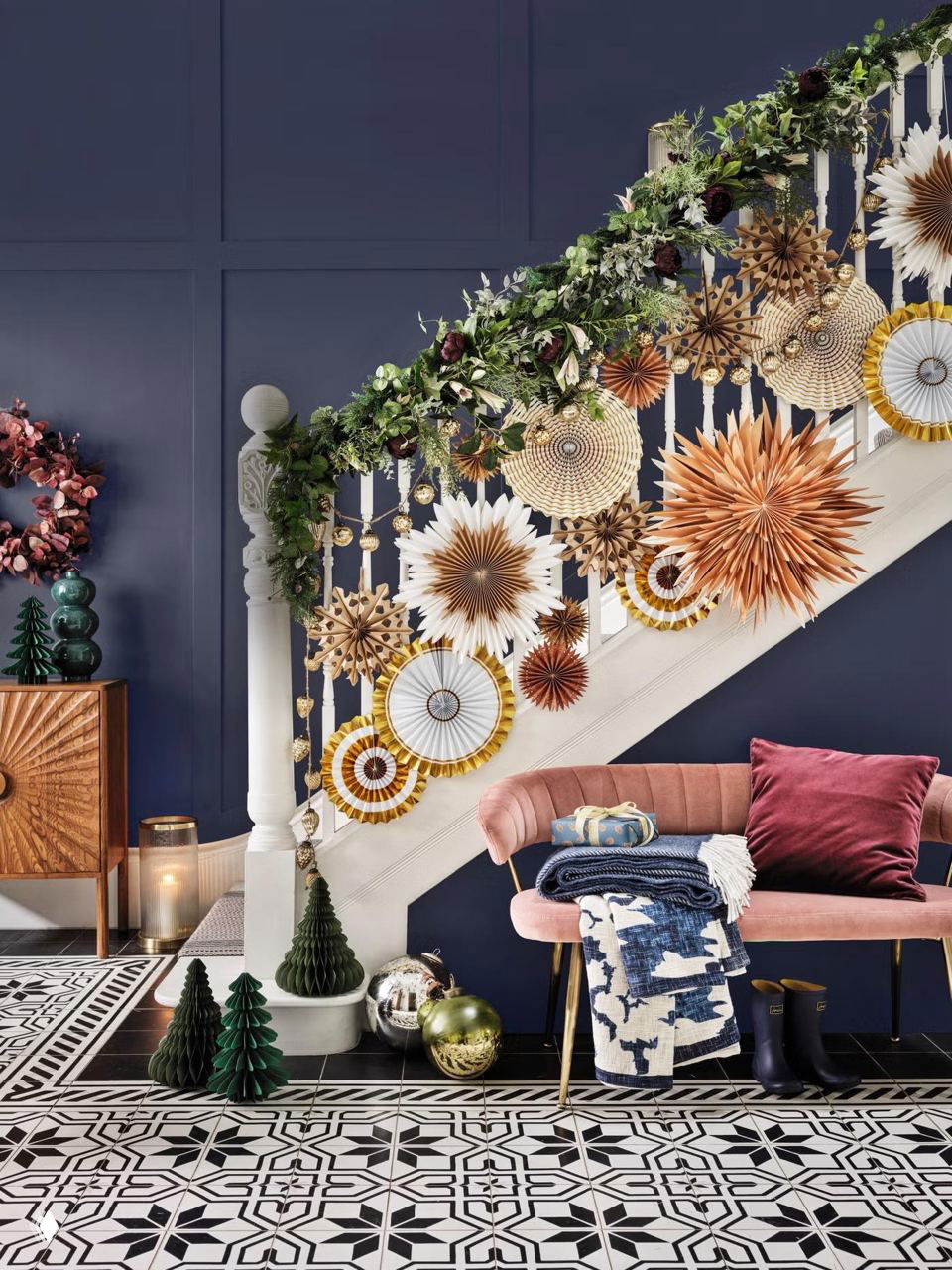 Лестничная ограда, украшенная зелёной гирляндой и бумажными звёздами в тёплых оттенках на фоне тёмной стены — декоративный акцент в коридоре.