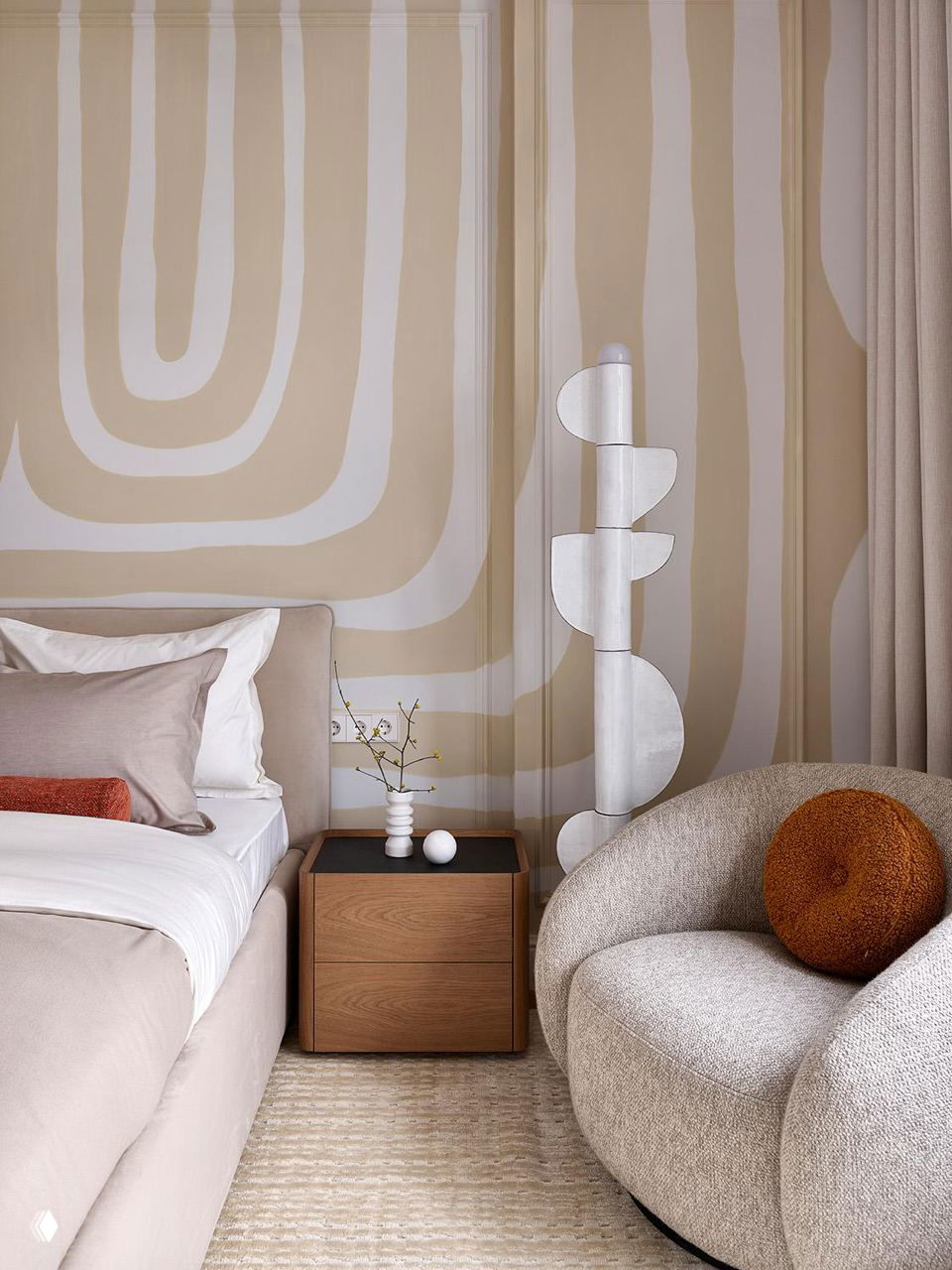 Спальня с мягкой мебелью в кремовой палитре, абстрактный орнамент на стене в спокойных тонах, деревянный прикроватный комод и округлое кресло в натуральных текстурах.