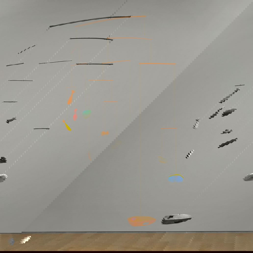 Крупный вид подвесного мобиля Колдера на серой стене: деревянные балки и разноцветные подвески разных форм создают воздушную кинетическую композицию.