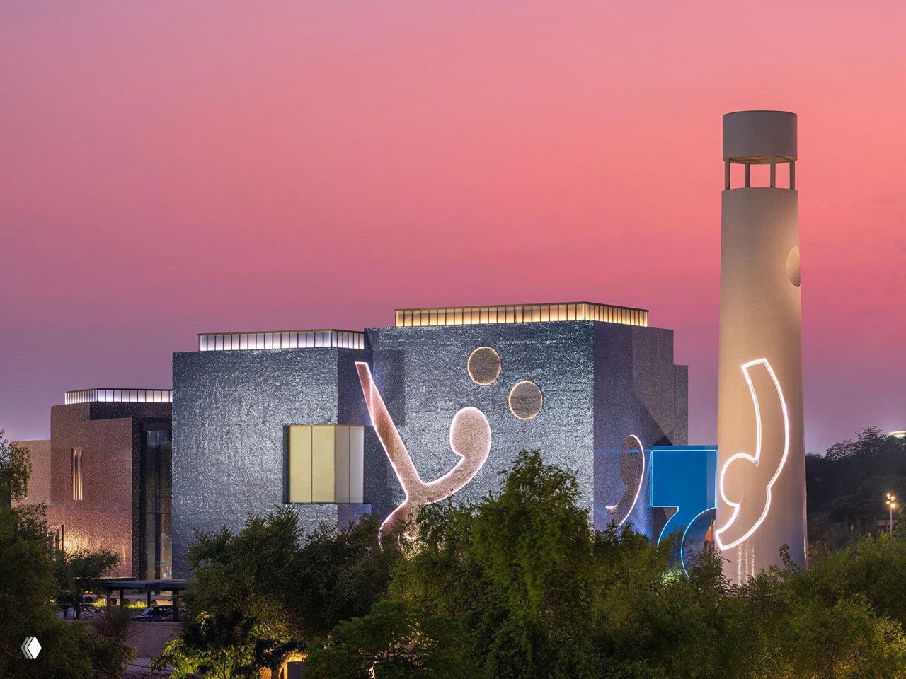 Фасад музея Макбула Фиды Хусейна в Дохе вечером: текстурная синяя стена с графическими символами и высокая цилиндрическая башня на фоне неба.