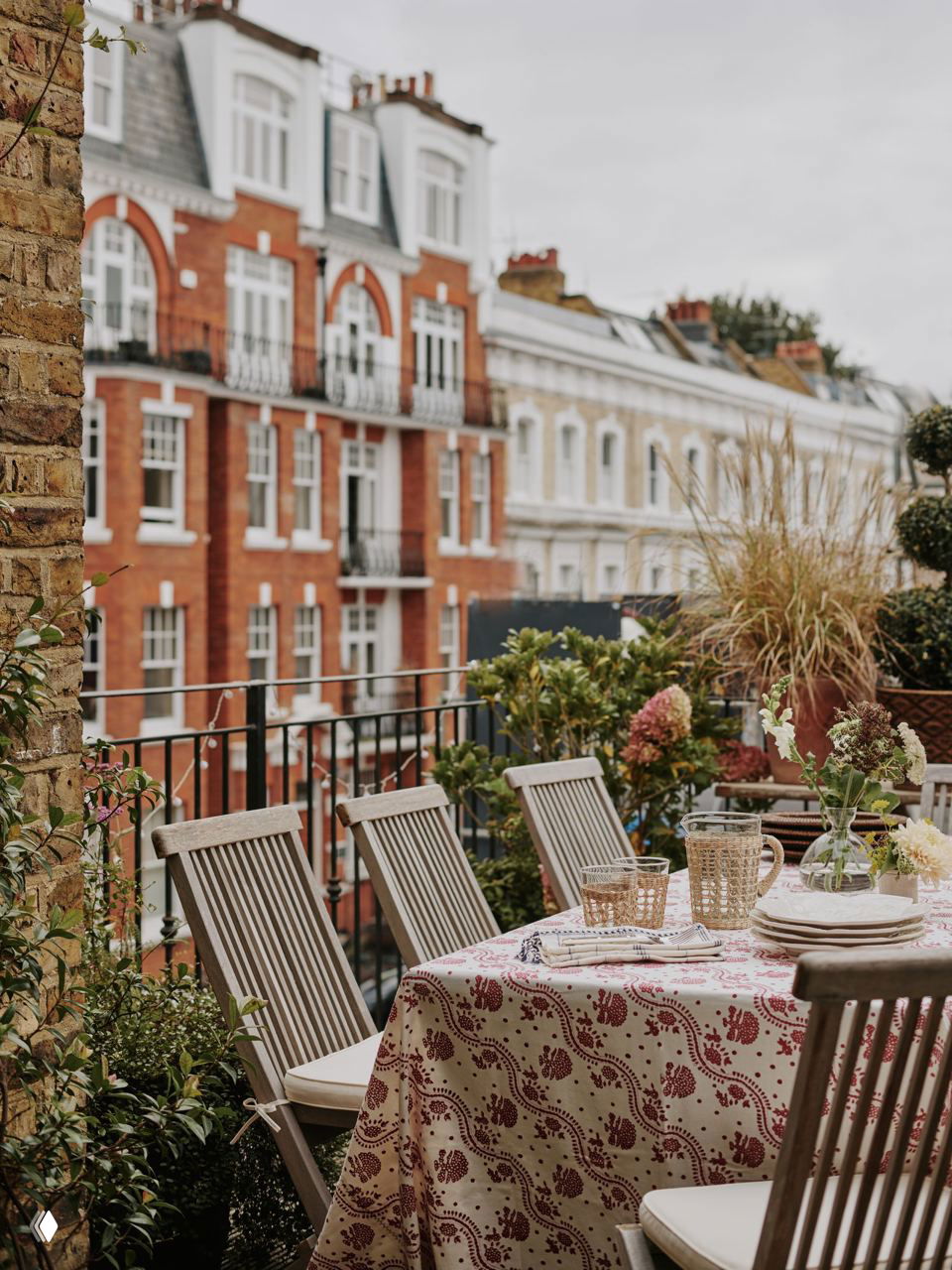 Балкон-терраса с обеденным столом и деревянными стульями, небольшими растениями и видом на кирпичные фасады викторианских домов района Челси в Лондоне.