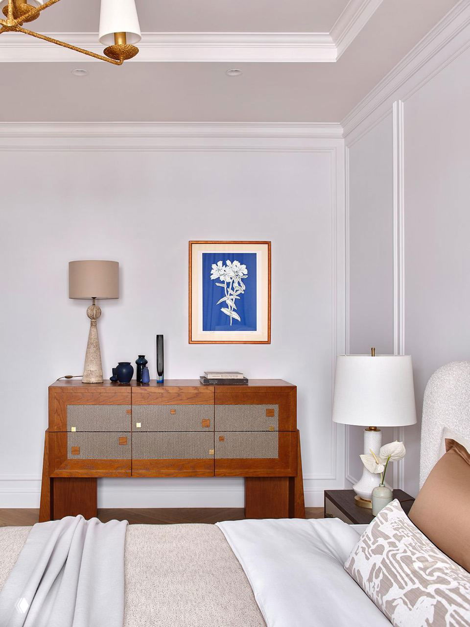 Деталь интерьера: винтажный деревянный комод, настольная лампа и компактное настенное панно на фоне светлой стены с классической лепниной.