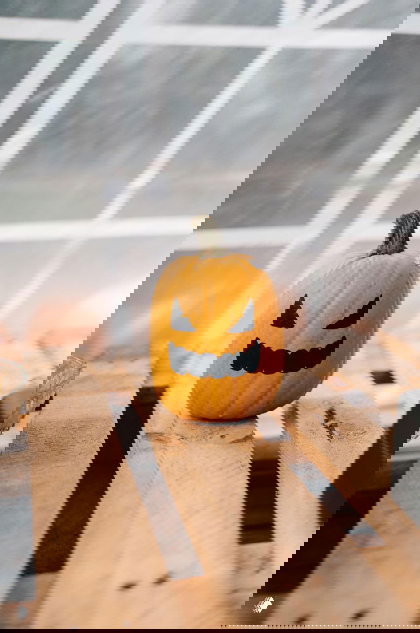 Небольшая тыква с зловещим нарисованным или вырезанным лицом, расположена на деревянной паллете у окна; дневной мягкий свет подчёркивает контур и текстуру кожуры.