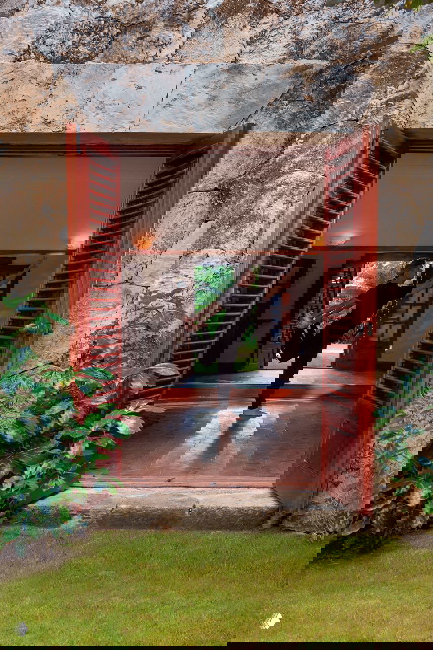 Открытая дверь с деревянными ставнями в доме в Доминикане; человек делает растяжку на фоне каменного портала и зелёного сада за входом.