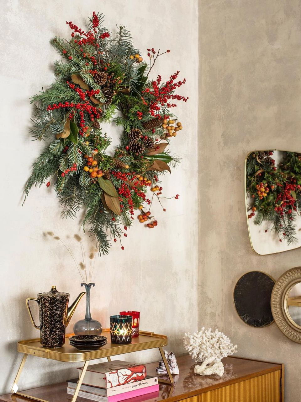 Пышный рождественский венок с ягодами и шишками на текстурной стене рядом с декоративным столиком, создающий элегантный праздничный уголок.