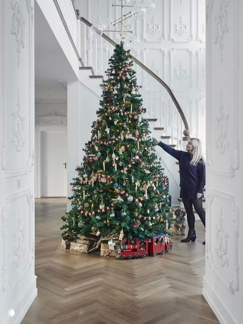 Высокая рождественская ёлка в светлом коридоре рядом со изящной лестницей; под ёлкой подарки и маленький игрушечный поезд, создающие атмосферу праздника.