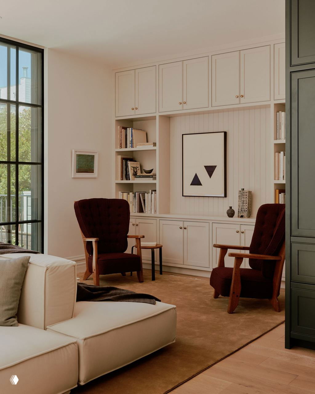 Уютная гостиная с мягким светлым диваном и двумя темно‑бордовыми креслами на фоне встроенных белых шкафов и полок с книгами и декором.