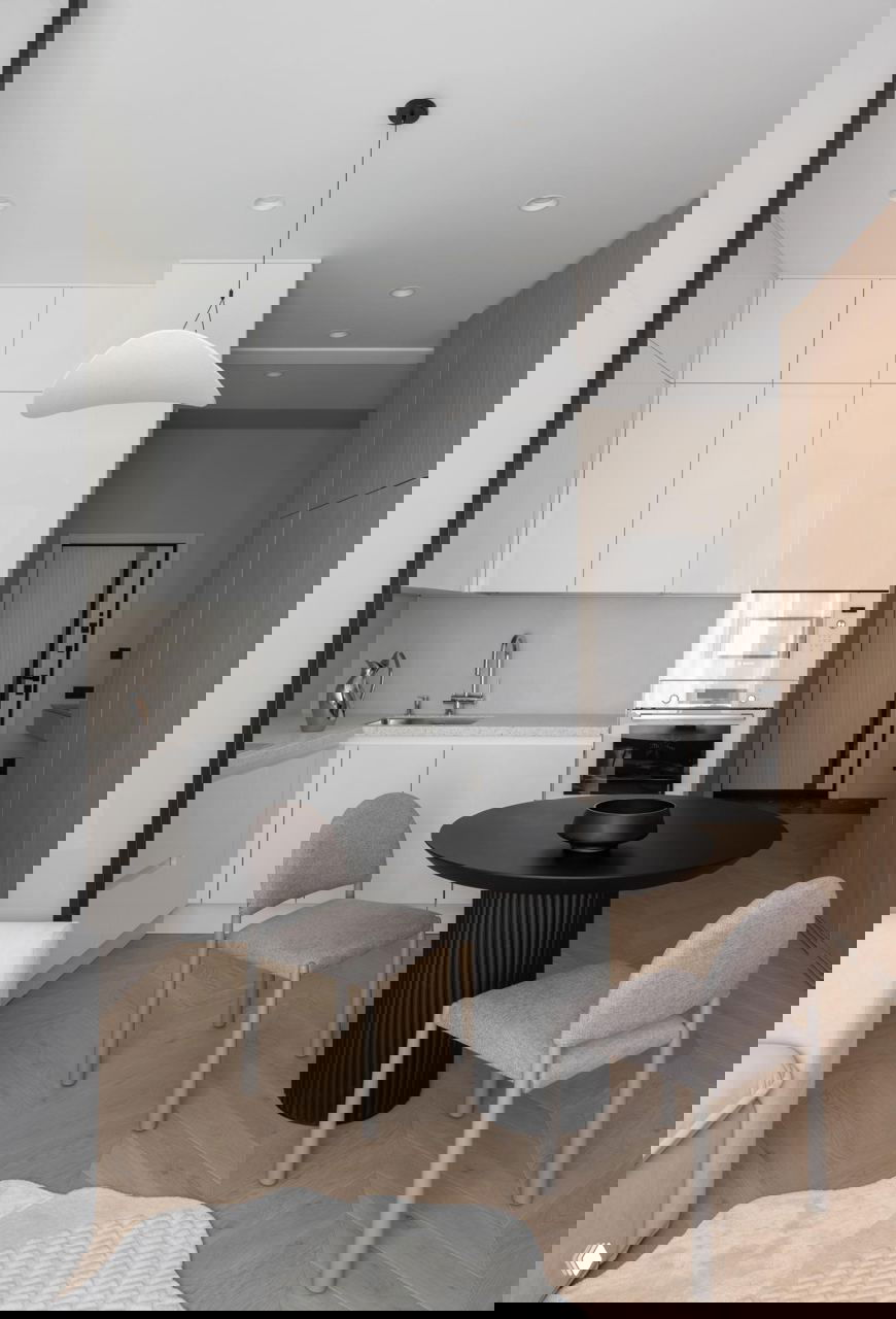 Вид на кухню и столовую: компактная белая кухня, чёрный стол и стулья, светлый паркет «ёлочкой» и нейтральная палитра материалов и фактур.