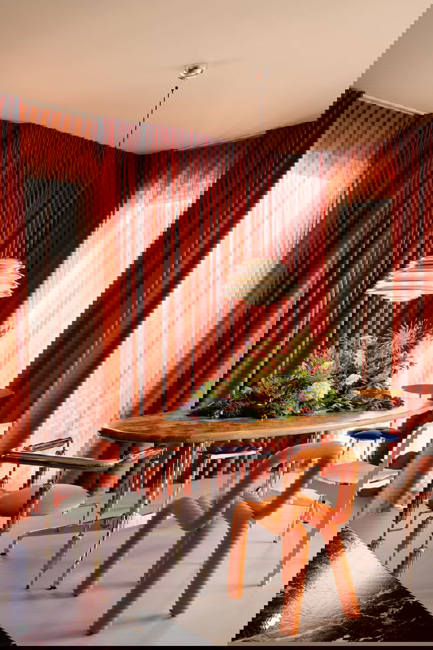 Гостиная-столовая с насыщенными терракотовыми шторами, овальным столом с цветочной композицией и эффектными дизайнерскими стульями в теплой палитре.