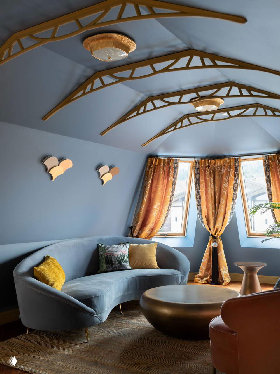 Гостиная на мансарде: голубая отделка потолка с декоративными балками, изогнутый синий диван и золотистые шторы — мягкое и камерное пространство.