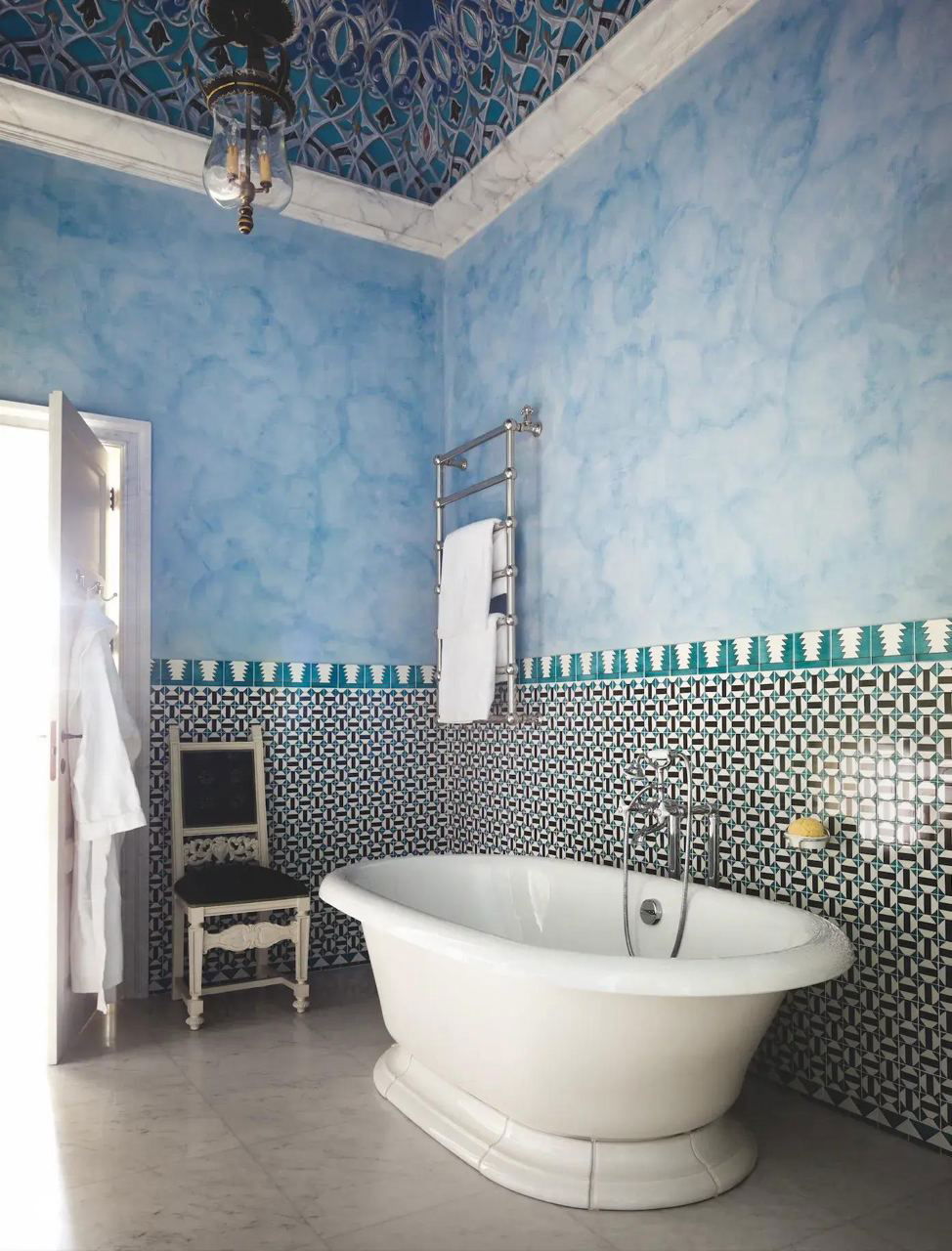 Ванная комната с отдельно стоящей белой ванной, настенным полотенцесушителем и декоративной плиткой с геометрическим узором, дополненная голубой фактурной штукатуркой.