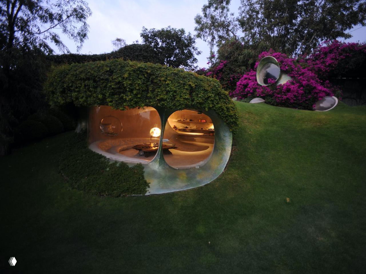Вечерний вид на встроенные в холм овальные помещения дома с подсветкой, из которых открывается вид на благоустроенный сад.