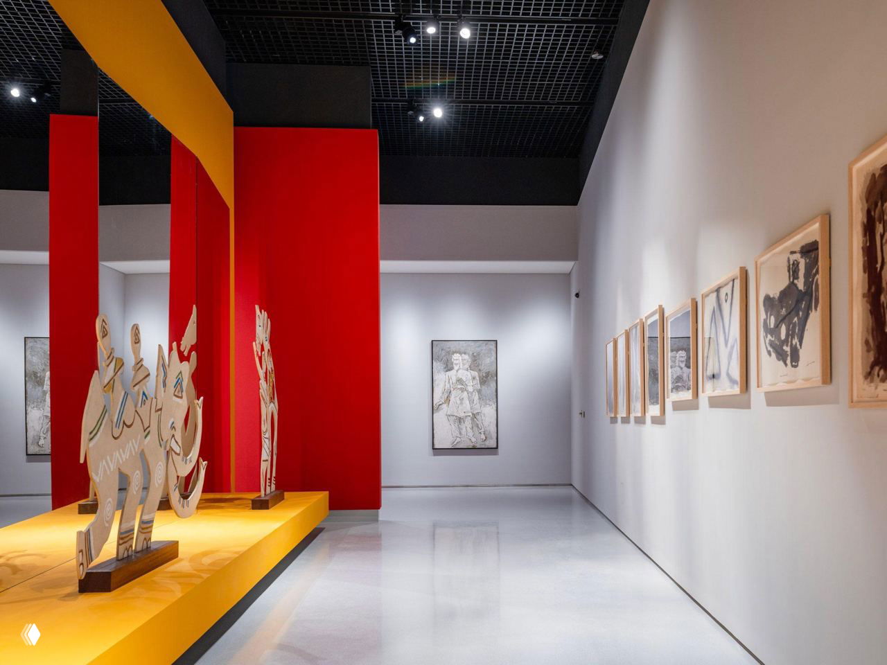 Зал с инсталляцией: ярко-жёлтая подиумная платформа, деревянные скульптуры и ряд работ на белой стене в музее Макбула Фиды Хусейна.