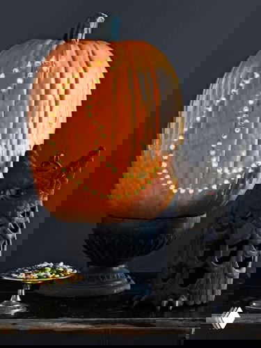 Высокая тыква с рисунком из множества отверстий в виде полумесяца, стоит на подставке рядом с декоративными элементами; контраст отверстий и подсветки создаёт узор.