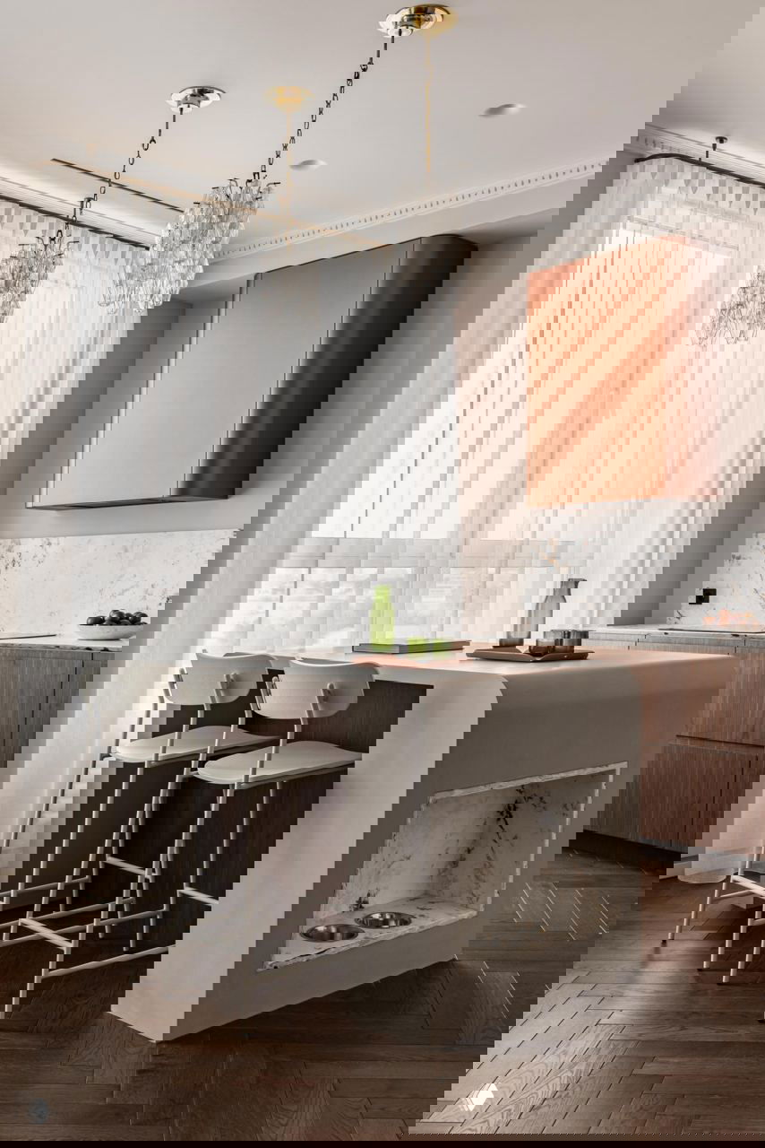 Кухня с барной стойкой и двумя высокими стульями, матовая фактура поверхности, мраморная столешница и большие светлые шторы на фоне окна.