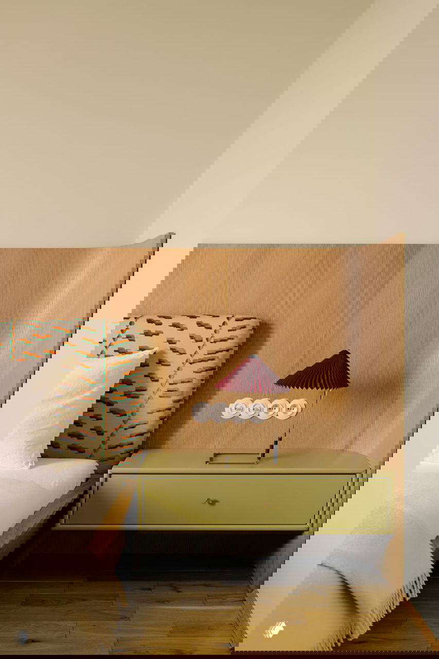 Спальня с деревянным изголовьем, прикроватной тумбой оливкового цвета и мягкой текстильной отделкой изголовья с принтом в спокойных тонах.