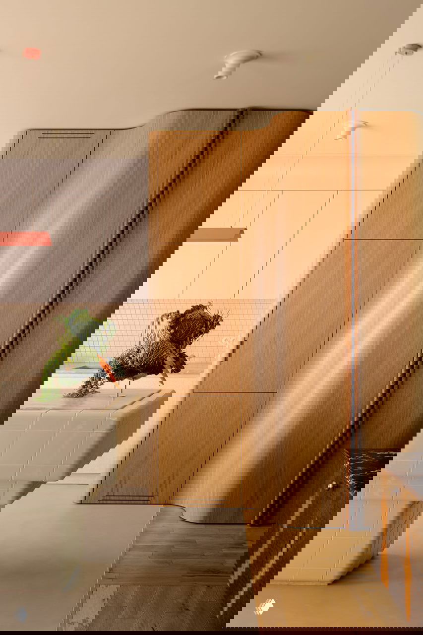 Фрагмент кухни: белые фасады, встроенные шкафы и деревянный изогнутый модуль рядом с плиточным островом на фоне нейтральных поверхностей.